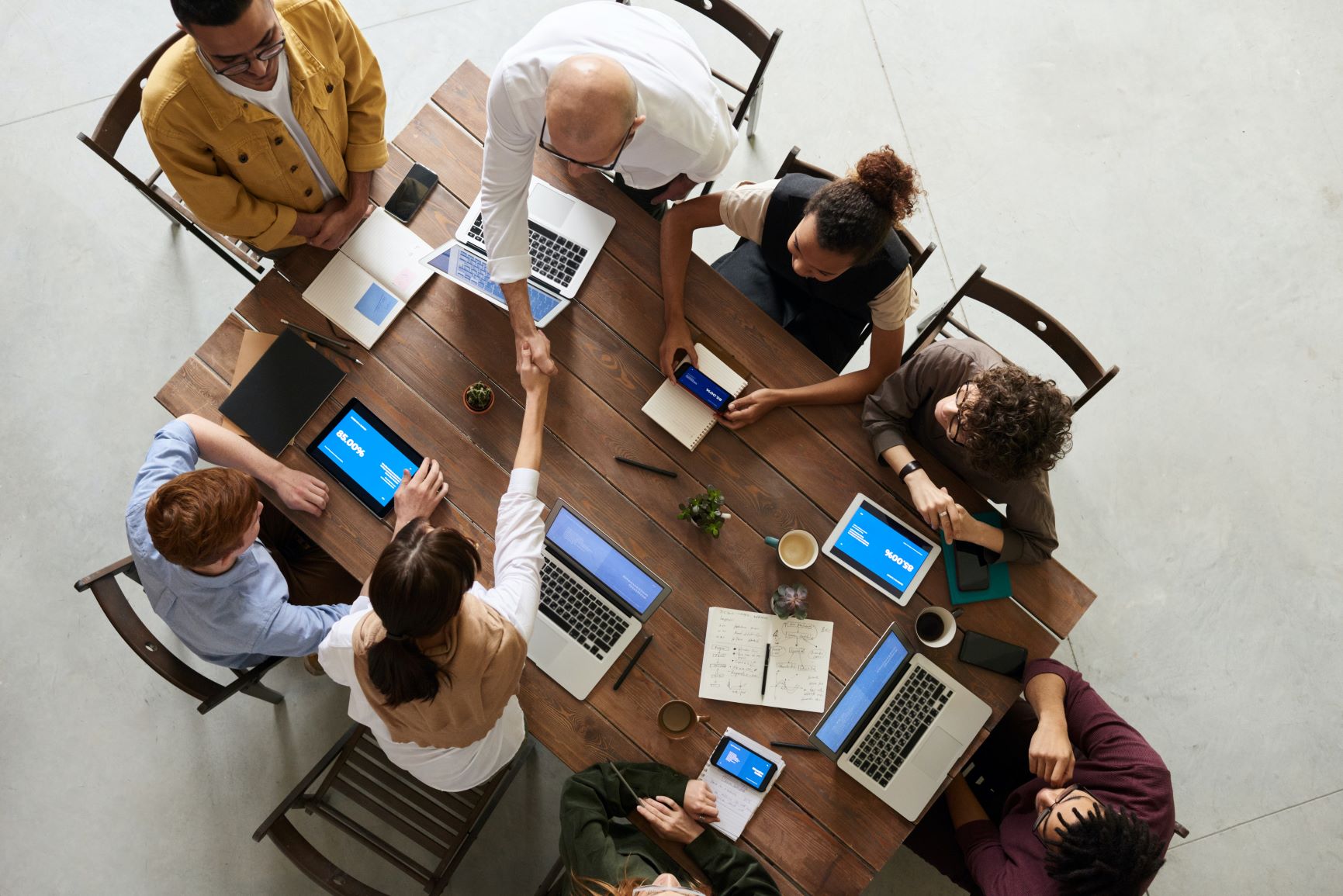 A group of professionals is having a meeting around a table with laptops and documents, engaging in a teamwork handshake.